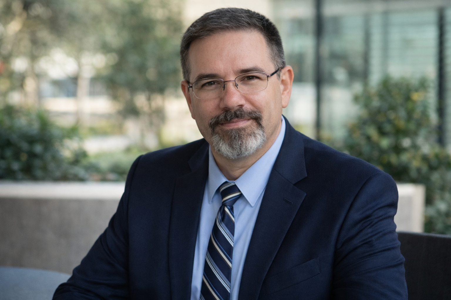 Professional headshot of Frank Jamison wearing a navy suit and striped tie, seated outdoors with a softly blurred background of greenery and modern architecture.