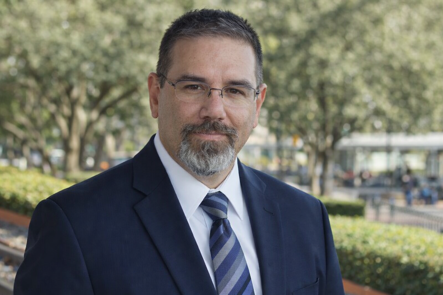Professional head and shoulders portrait of Frank Jamison wearing a navy suit, white dress shirt, and striped blue tie, standing outdoors with a softly blurred park background and looking directly at the camera with a confident expression.