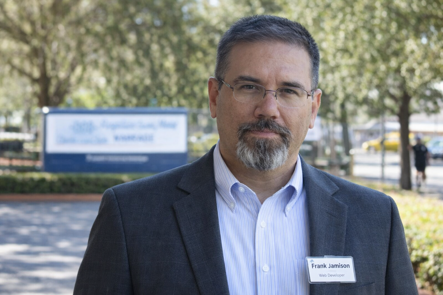 Frank Jamison standing outdoors in professional attire, wearing a charcoal blazer and striped dress shirt, facing the camera with a confident expression, trees and a campus style sign softly blurred in the background.